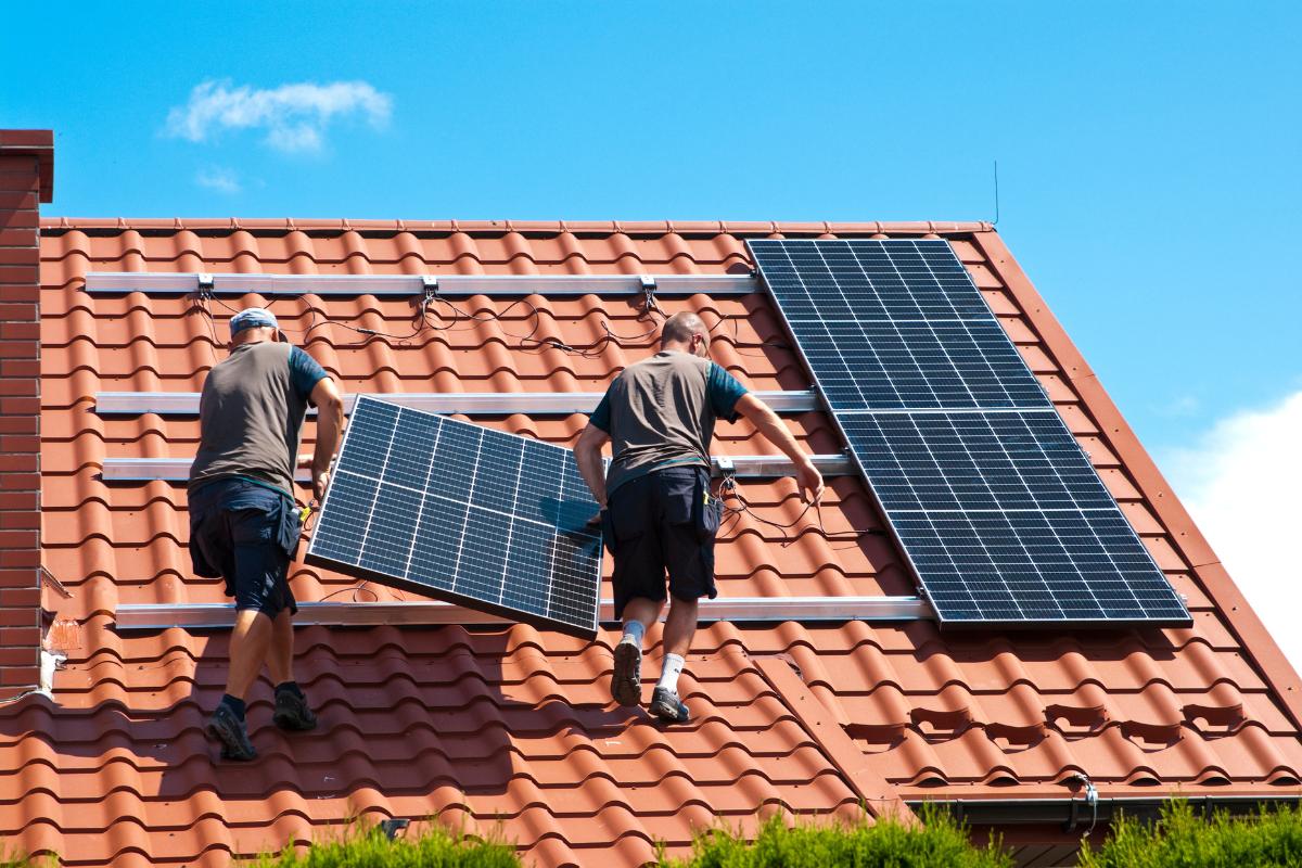 Zwei Männer tragen ein Solarpanel zur Unterkonstruktion auf dem Dach. Zwei Solarpanele sind schon montiert. 