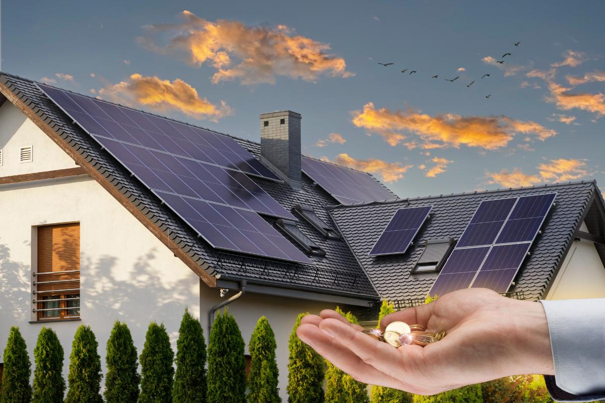 Im Hintergrund steht ein Haus mit Solarpanelen auf dem Dach, im Vordergrund ist eine Hand mit Münzen in der Handfläche zu sehen.